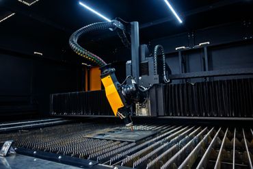 Industrielle Laserschneidanlage in einer dunklen Fabrikhalle, mit einem leuchtend gelben Arm über einem Metallarbeitstisch