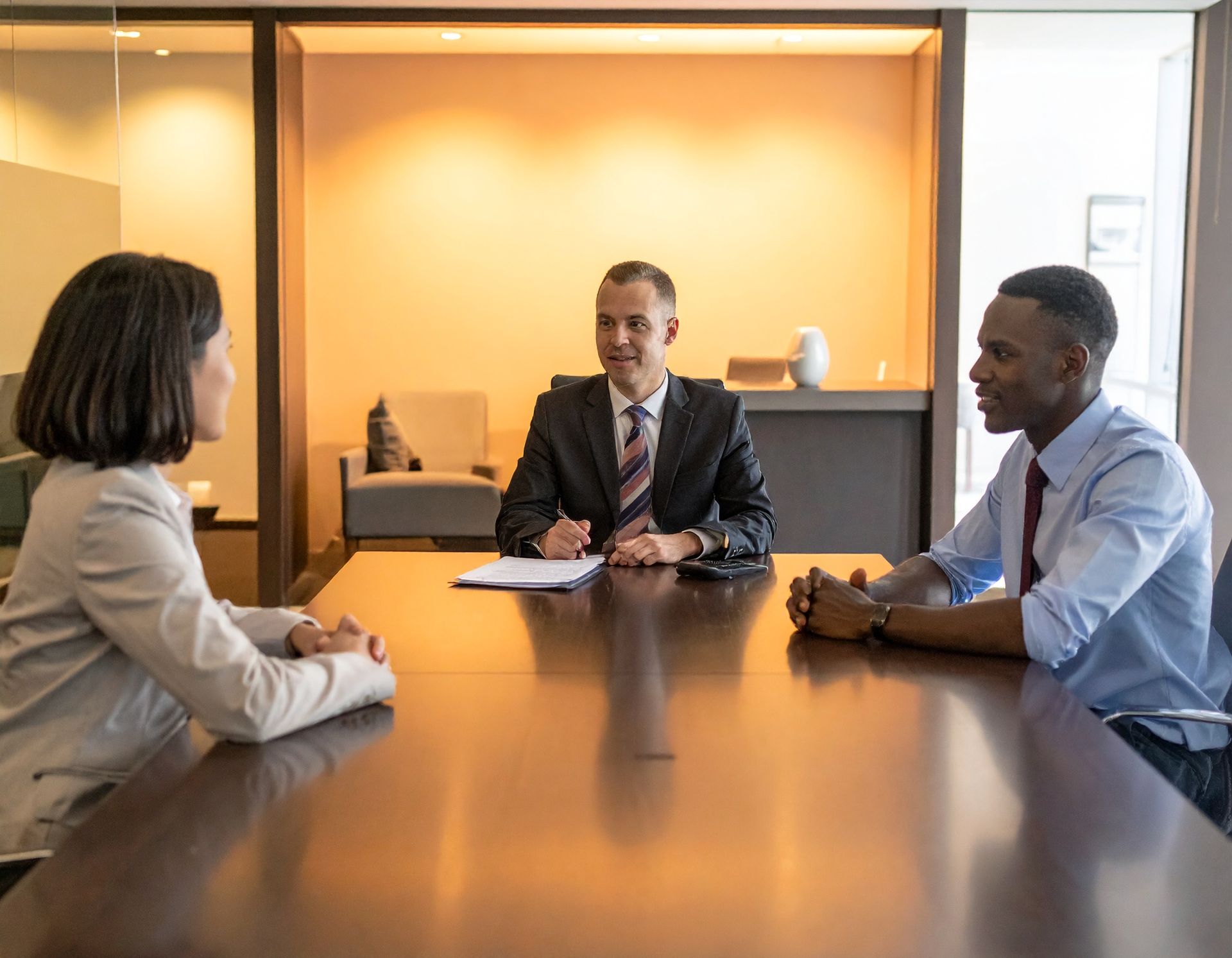 Three people in business attire seated around a conference table in a meeting room.