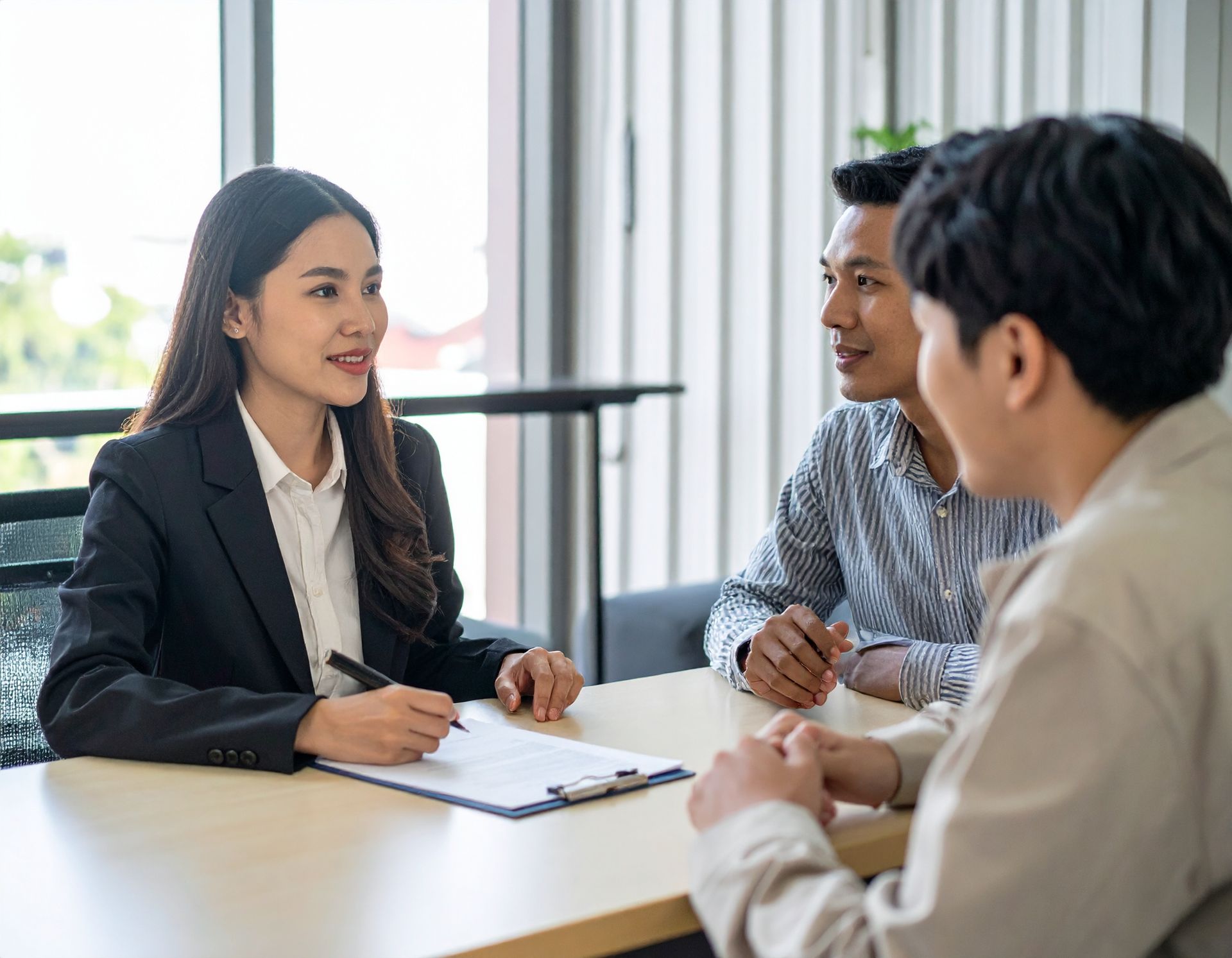 Three people in a bright office meeting, discussing around a table with a notebook.