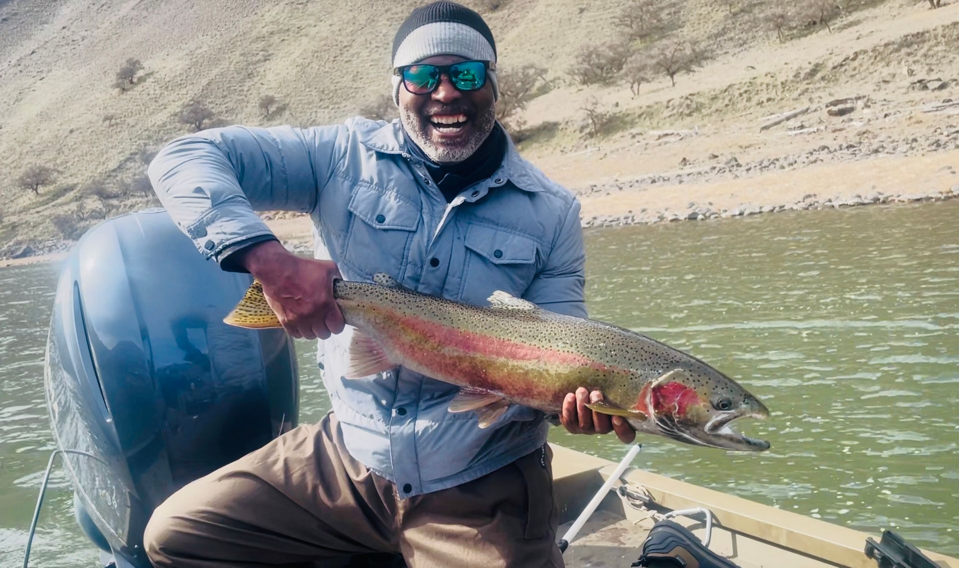 Man on a boat, holding a large rainbow trout. Smiling with a mountain backdrop.