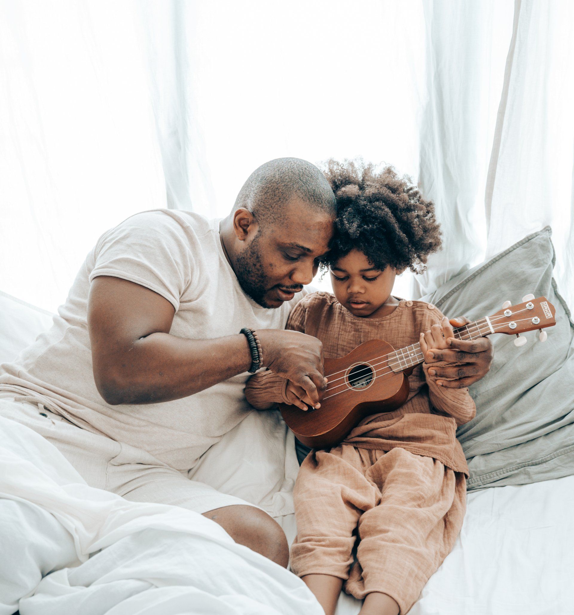 Man teaching a child to play ukulele, both sitting on a bed.
