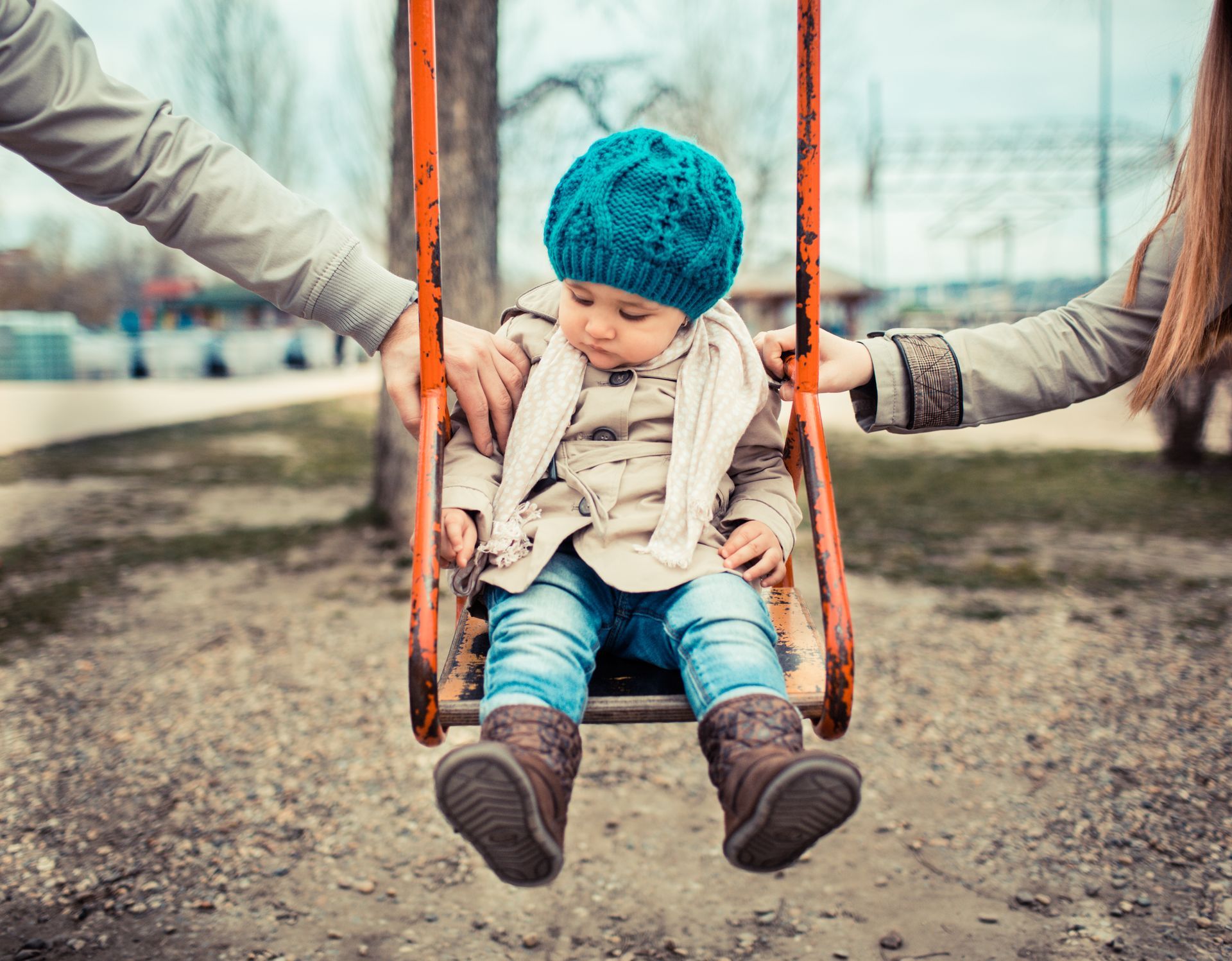 Child on a swing between two adults, hands on swing. Overcast day.