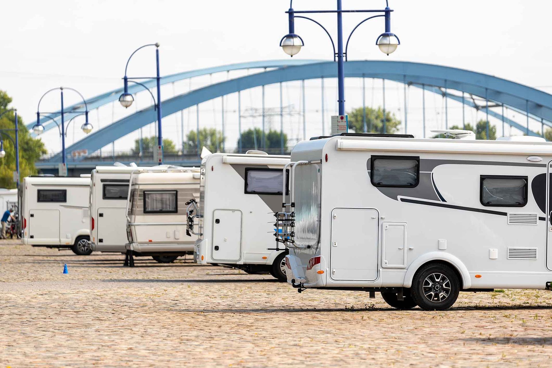 Four white RVs parked on gravel, with a blue arch bridge in the background.