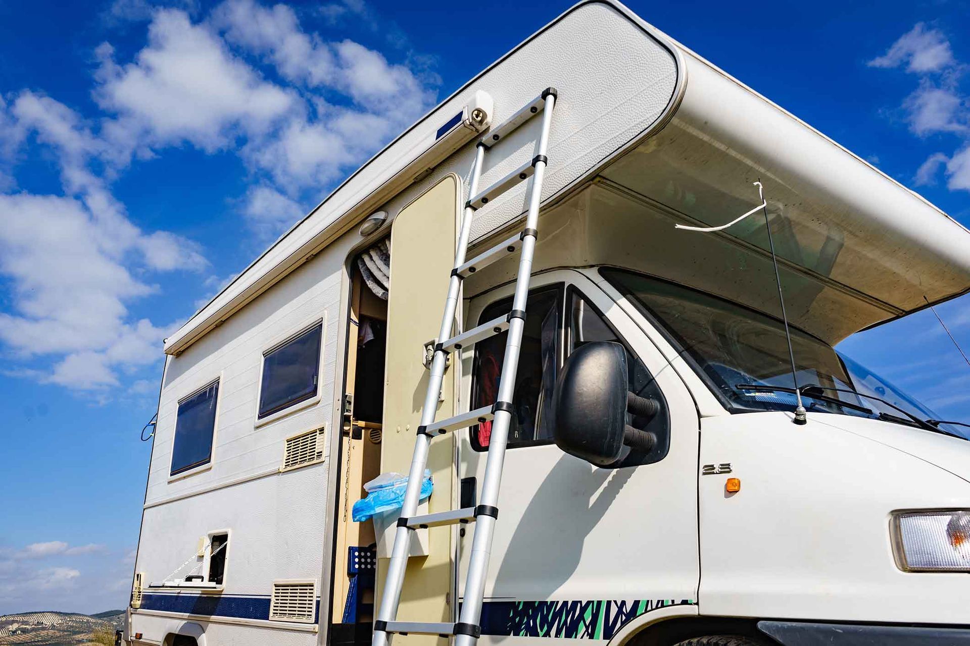White RV with ladder extended, open door, and blue sky background.