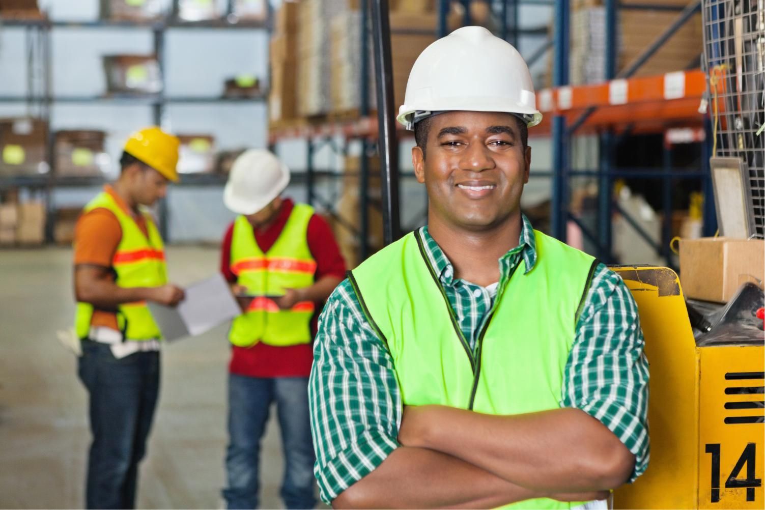 Warehouse worker in white hard hat and green vest stands with arms crossed; coworkers work in background.