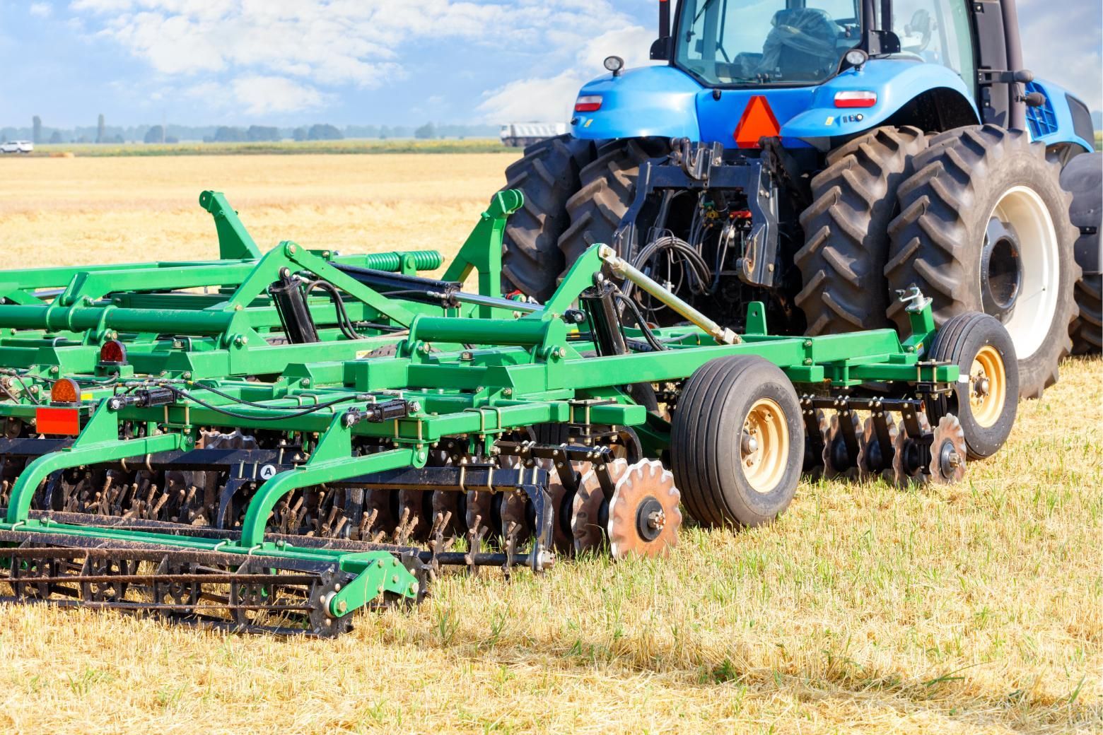 Blue tractor pulling a green disc harrow across a harvested field under a bright sky