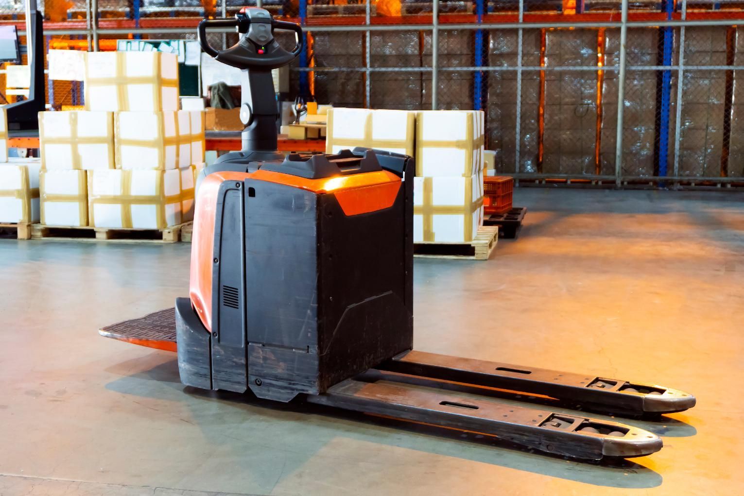 Orange and black warehouse pallet jack parked on a warehouse floor near stacked boxes.