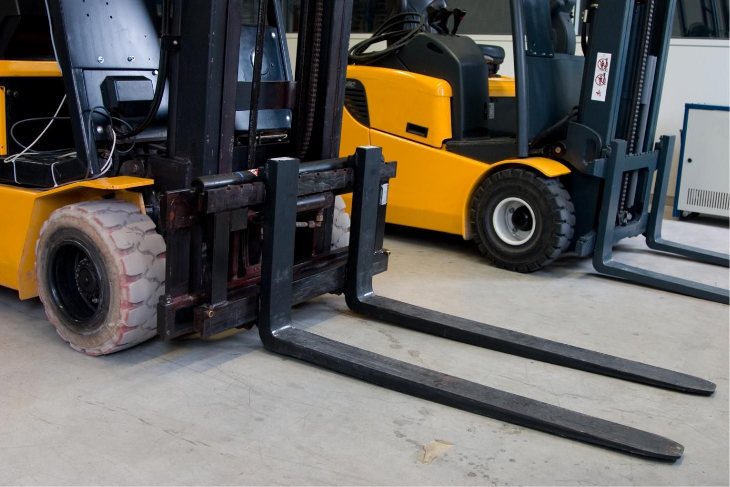 Two yellow forklifts parked side by side, one with long black forks extended on a warehouse floor