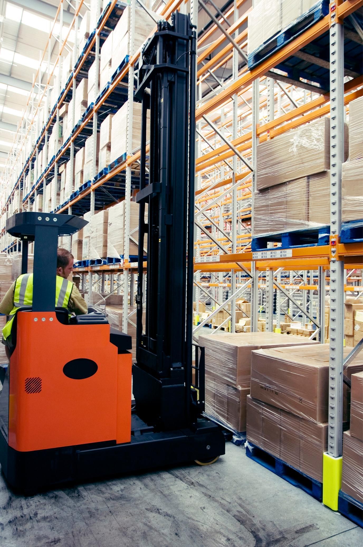 Orange warehouse forklift moving down an aisle beside tall pallet racks and stacked boxes