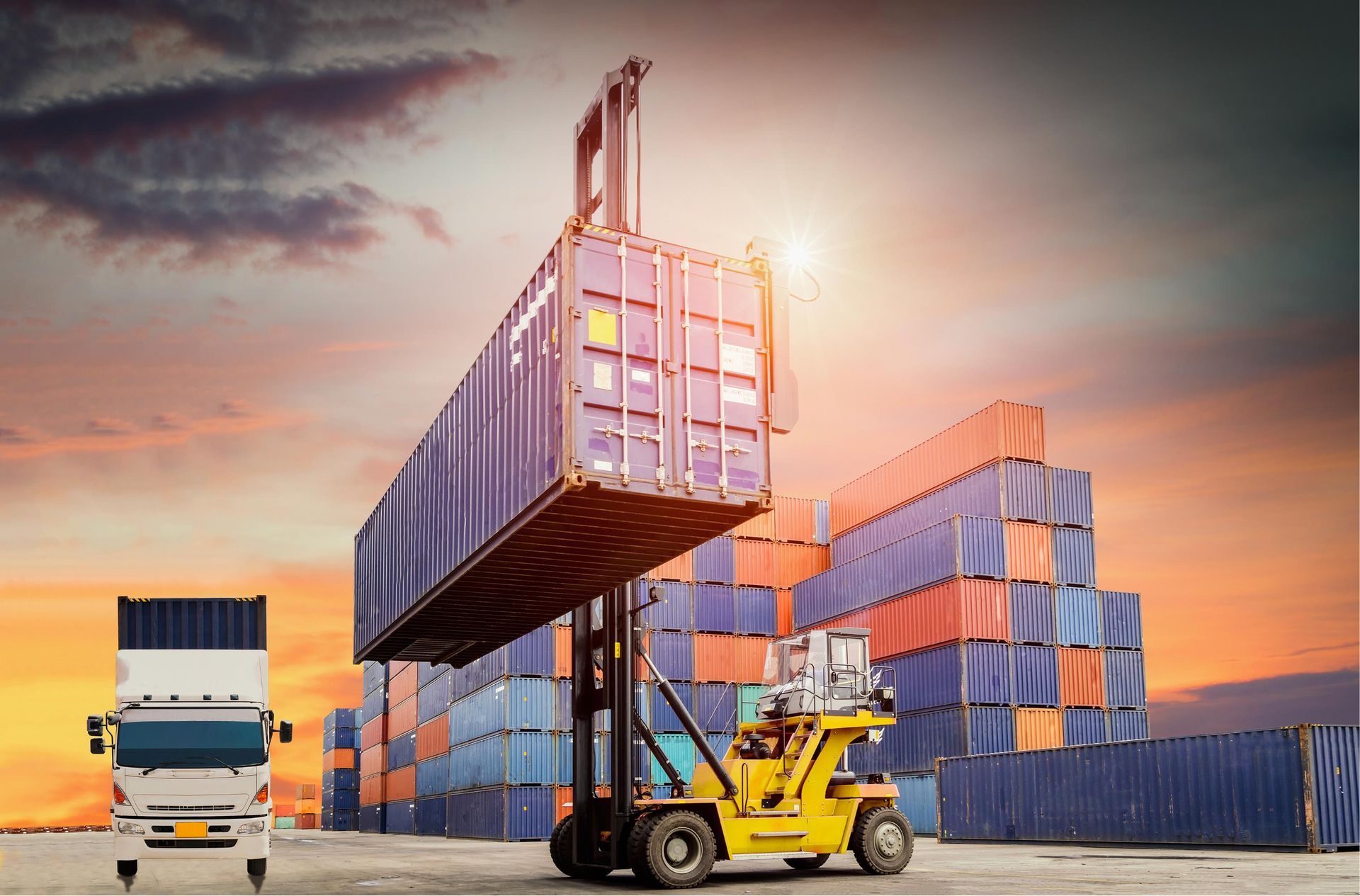 Forklift lifting shipping containers in a port at sunset, with a truck and stacked containers nearby