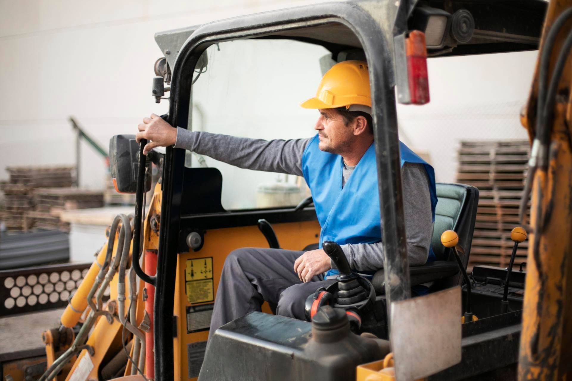 Worker in yellow hard hat operating a forklift in a warehouse, with pallets stacked nearby.