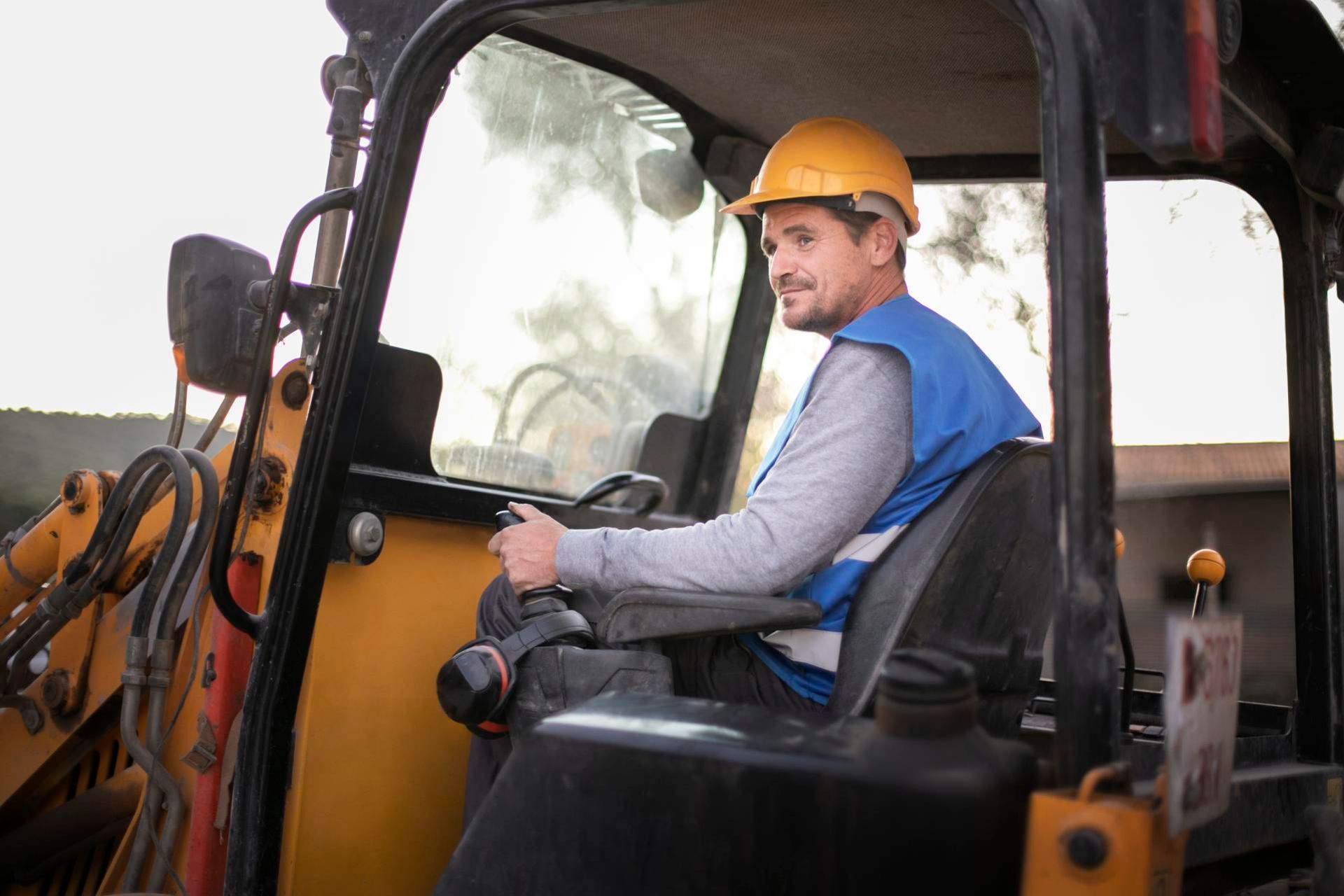 Construction worker in yellow hard hat operating a backhoe inside the cab