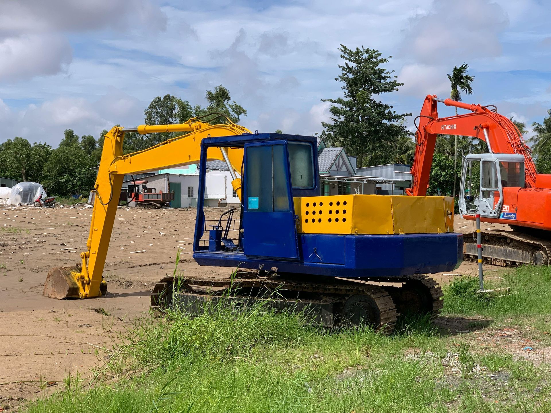 Yellow and blue excavator on dirt lot with orange excavator in the background