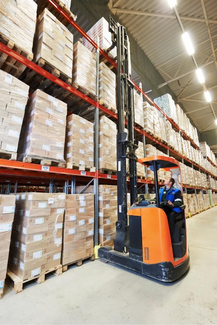 Worker driving a forklift in a warehouse aisle beside tall stacks of boxed pallets