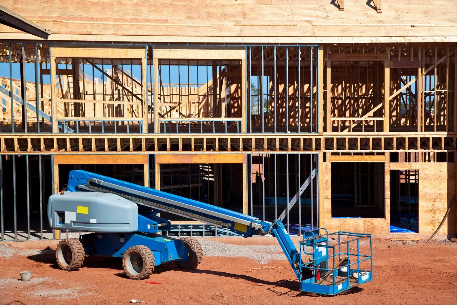 Blue boom lift at a wooden house under construction, with framing exposed and scaffold-like rails above.