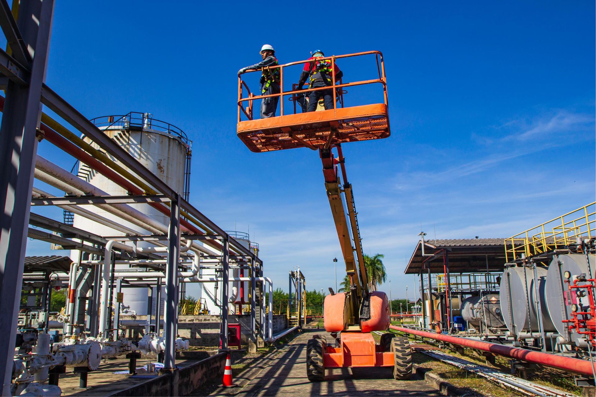 Workers in an orange lift inspecting industrial piping at a refinery under a bright blue sky