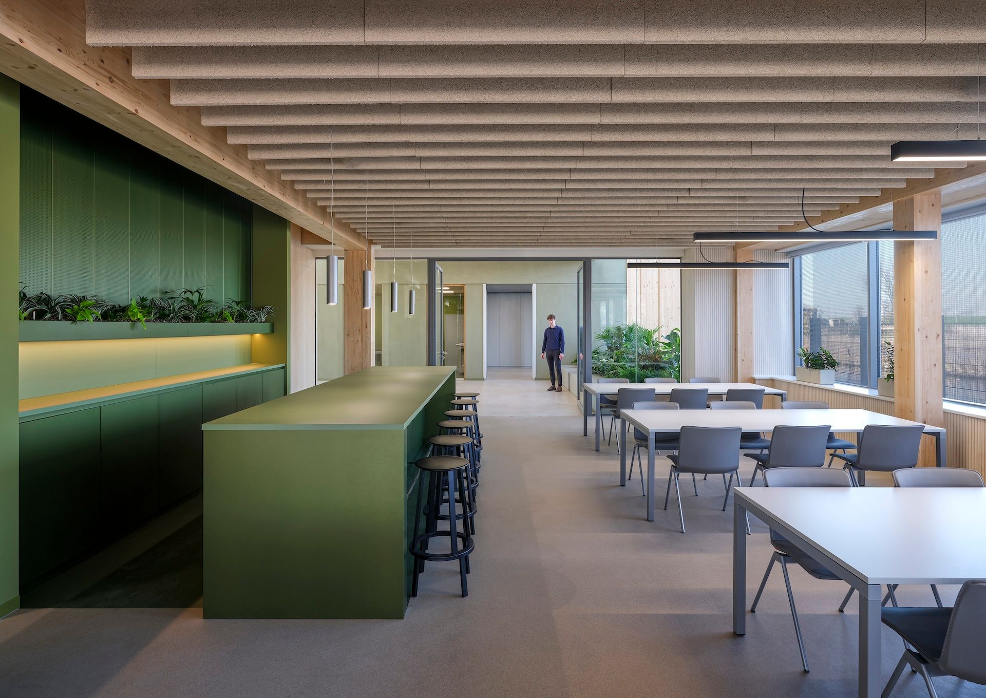 Modern office lounge with green bar, tables, chairs, and a person walking in the background