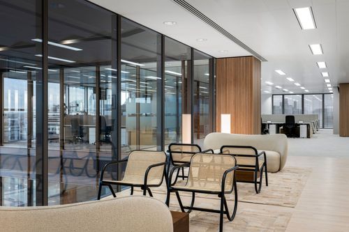 Modern office lounge with glass walls, woven chairs, beige sofas, and bright overhead lighting