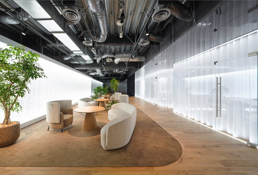 Modern office lounge with round table, curved chairs, indoor tree, wood floor, and glass wall corridor