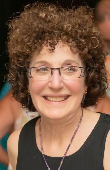 Smiling person with curly hair and glasses wearing a black top and purple necklace