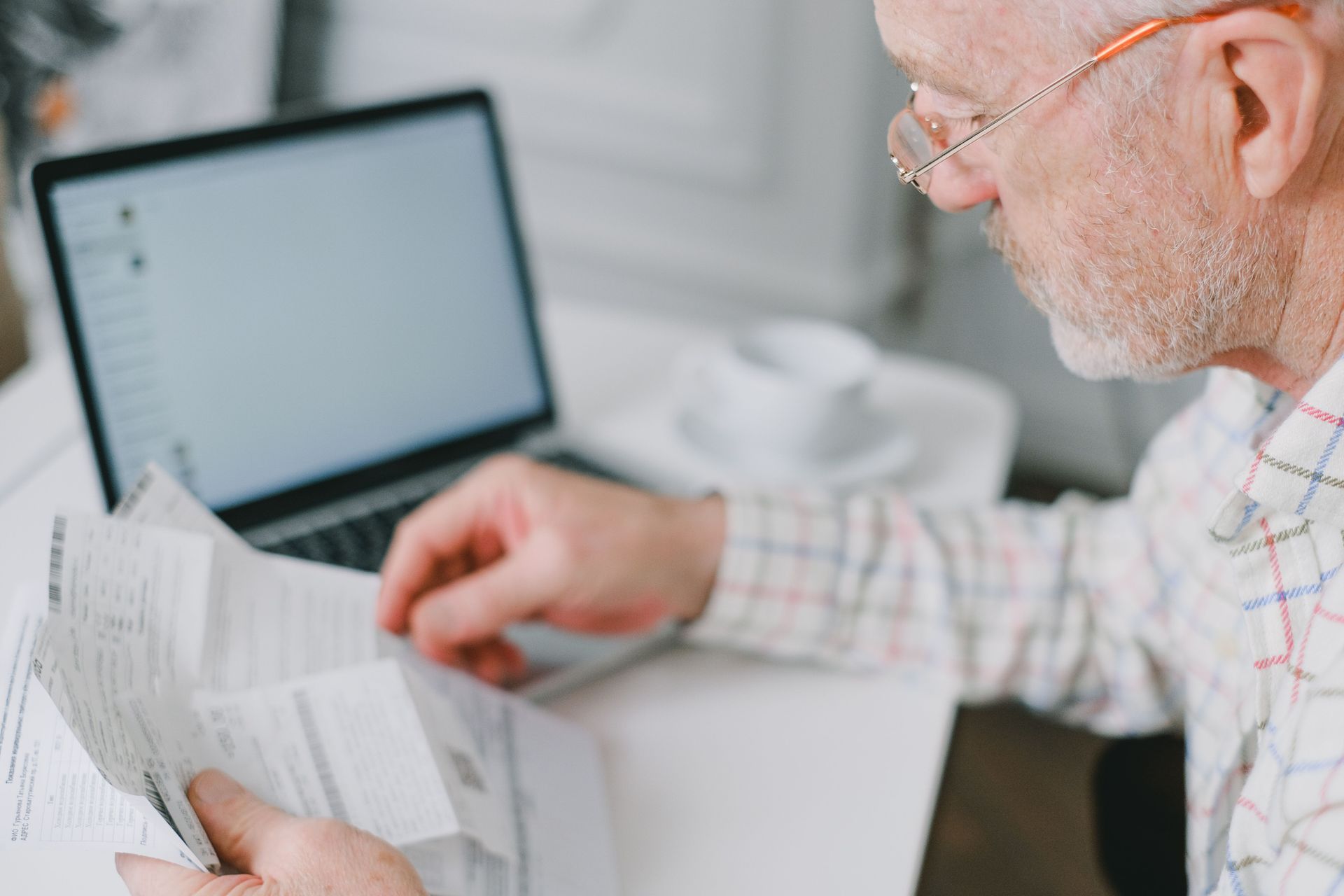 Man reading papers beside a laptop on a desk, in a bright home office