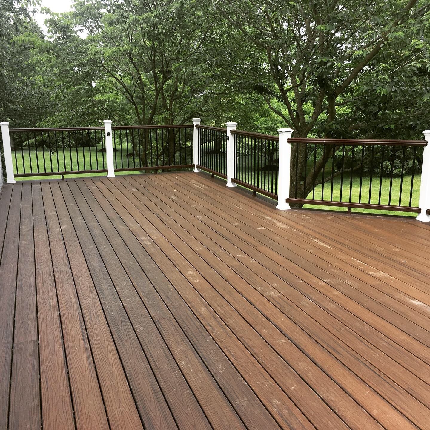 Wooden deck with black railing and white posts, surrounded by green trees.