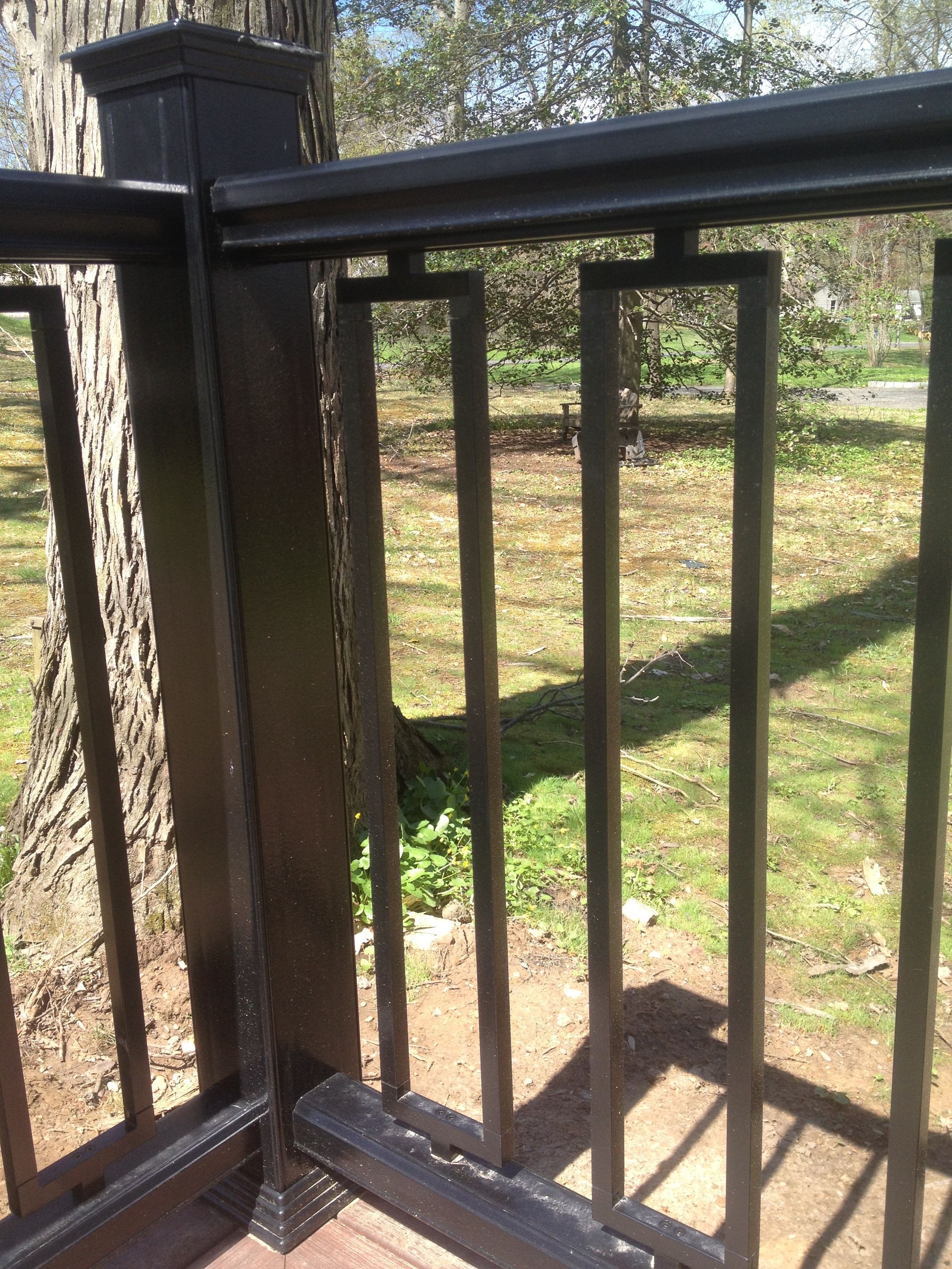 Black deck railing with vertical square balusters against a blurred green and brown outdoor setting.