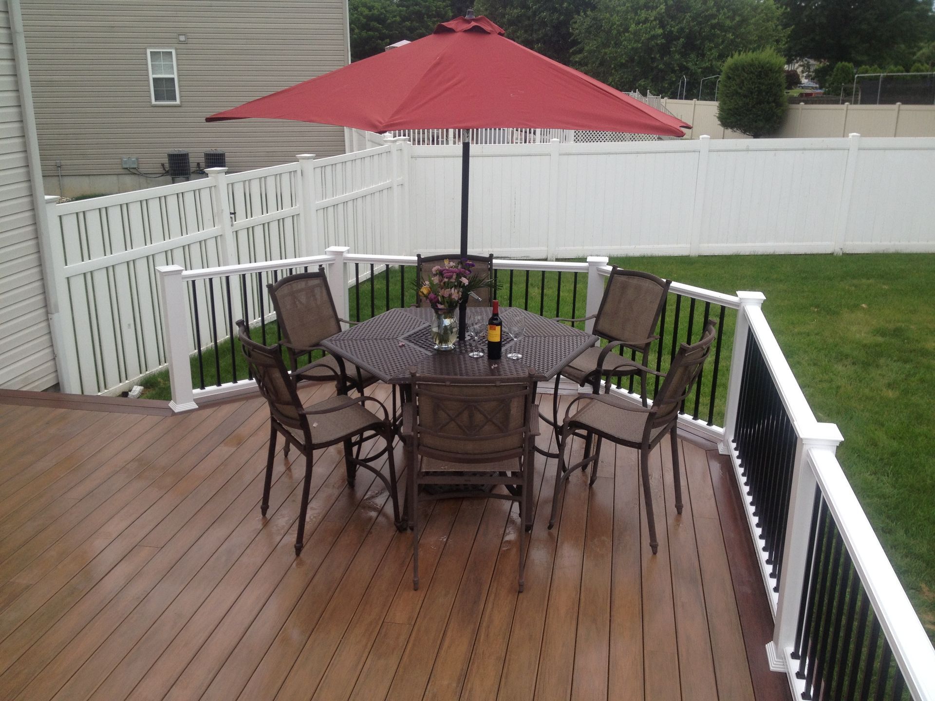 Deck with table, chairs, and red umbrella surrounded by a white fence and green lawn.