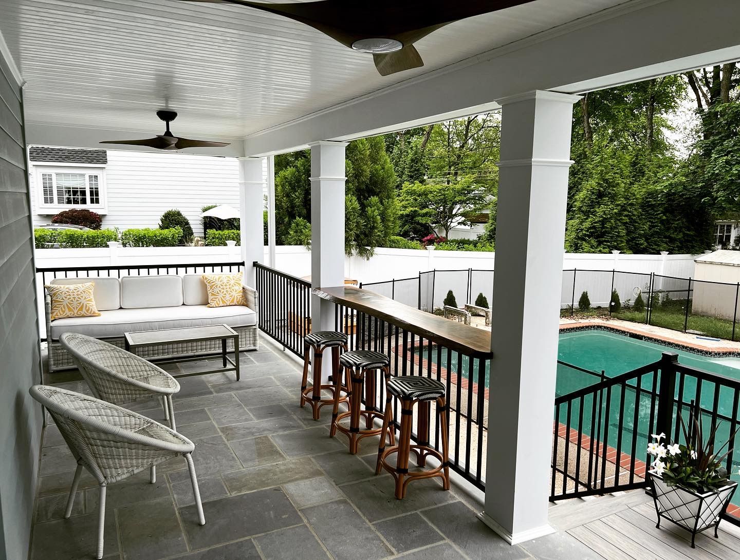 Patio with bar and pool view; wicker furniture, gray stone floor, white pillars, black railings.