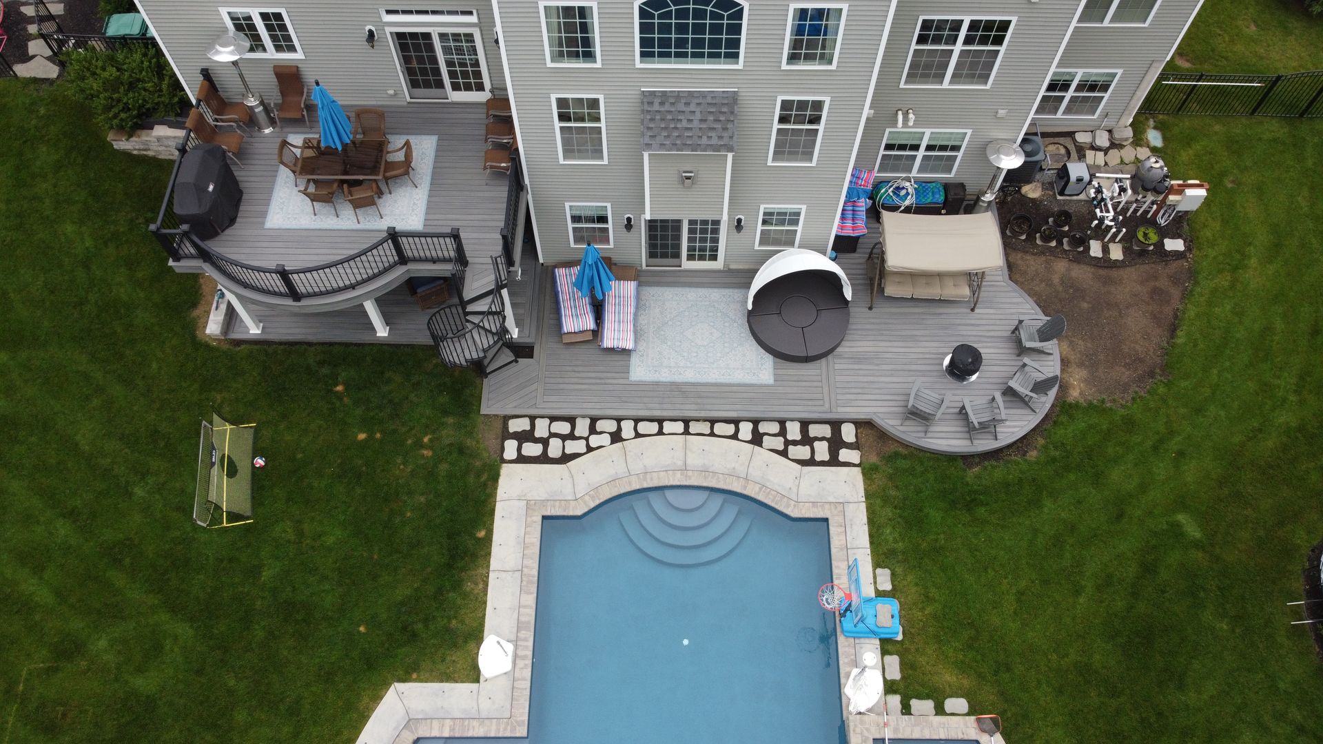 Aerial view of a house with a pool, multiple decks, and a green lawn. Blue pool and decks, grey house.