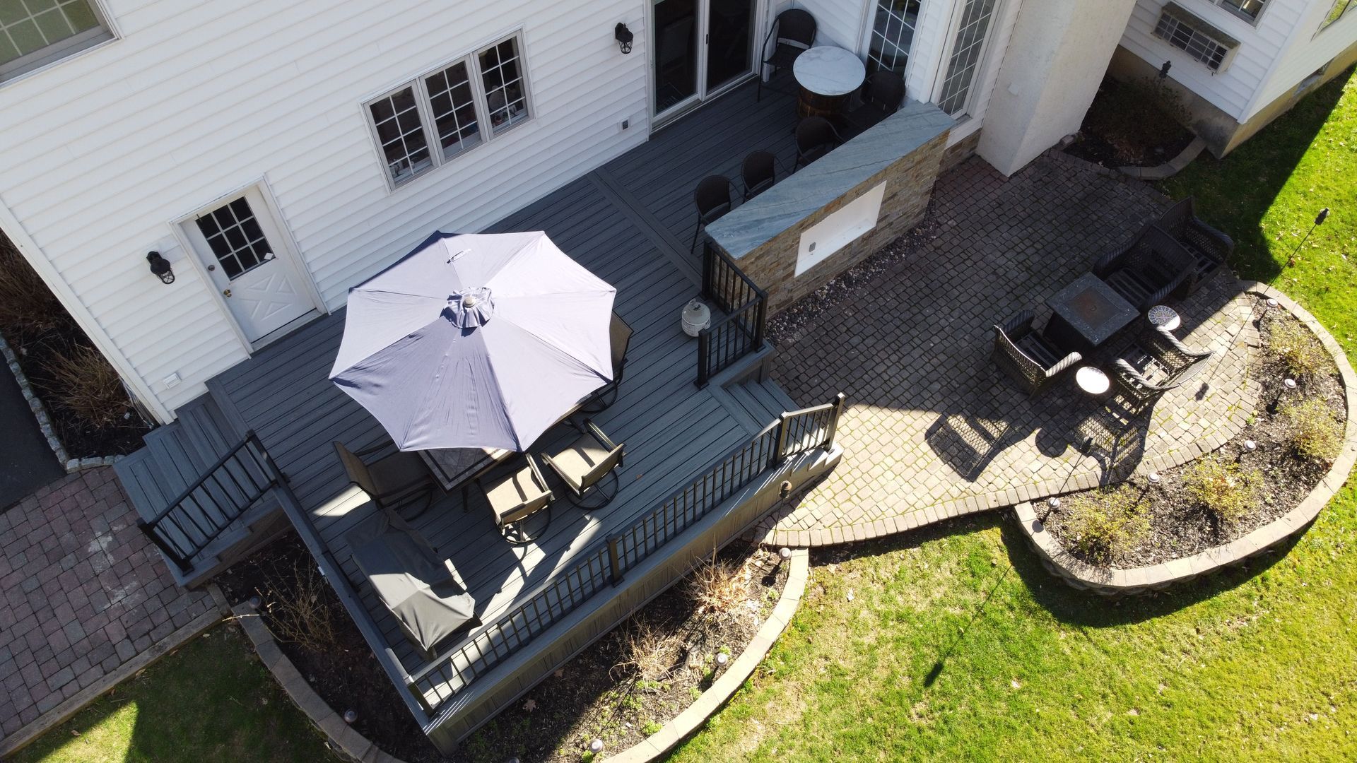 Overhead view of a deck with an umbrella, seating, a bar, and a brick patio in a backyard with green grass.