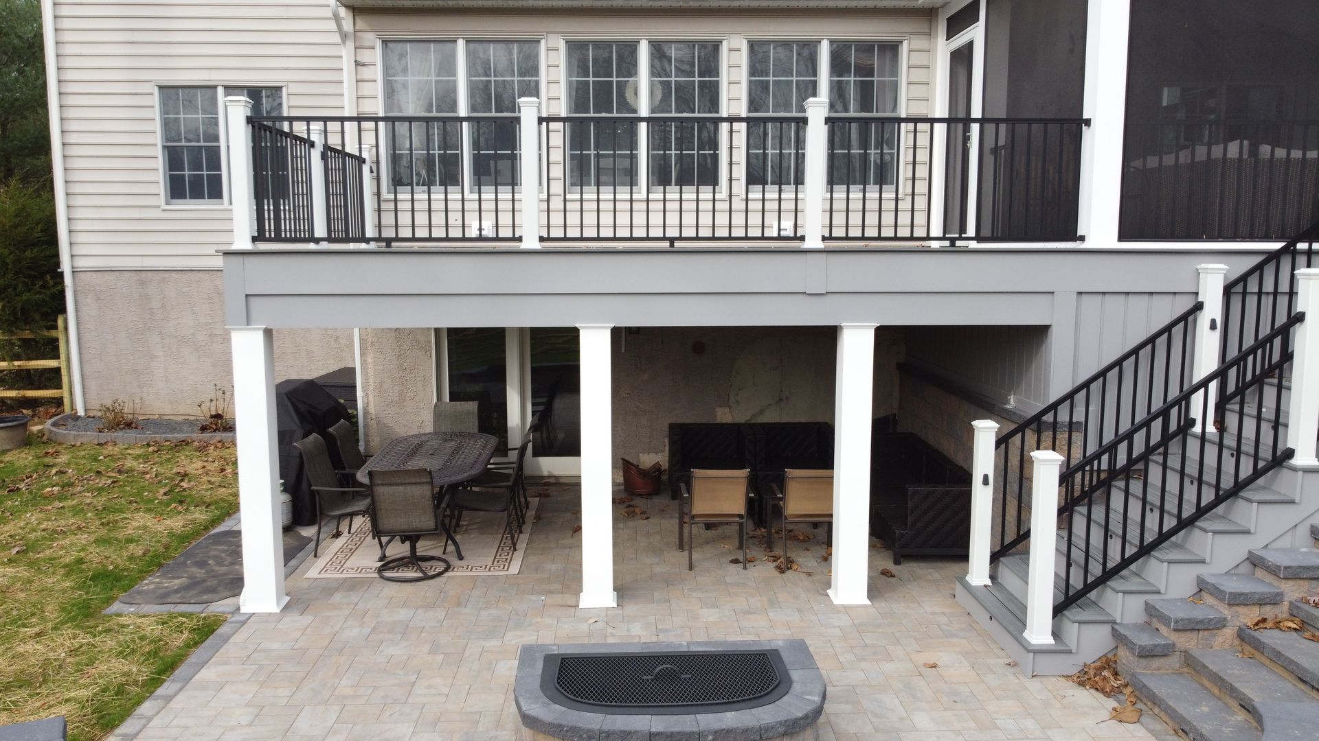 Deck with black railing, white columns, and patio furniture under a covered area. Gray brick patio with fire pit.