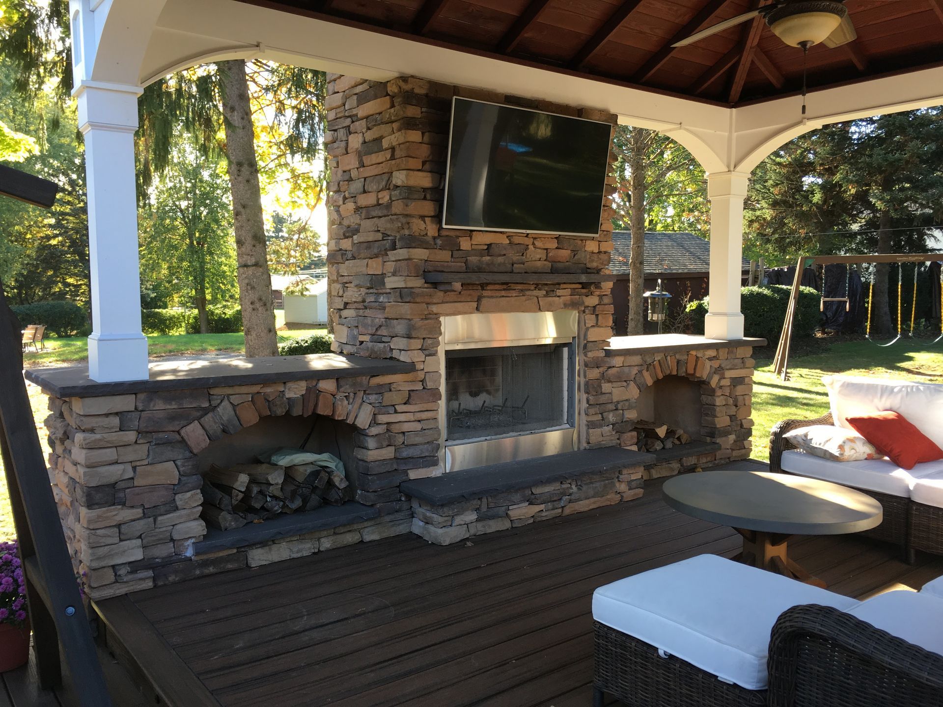 Stone outdoor fireplace with a television and seating under a gazebo roof.