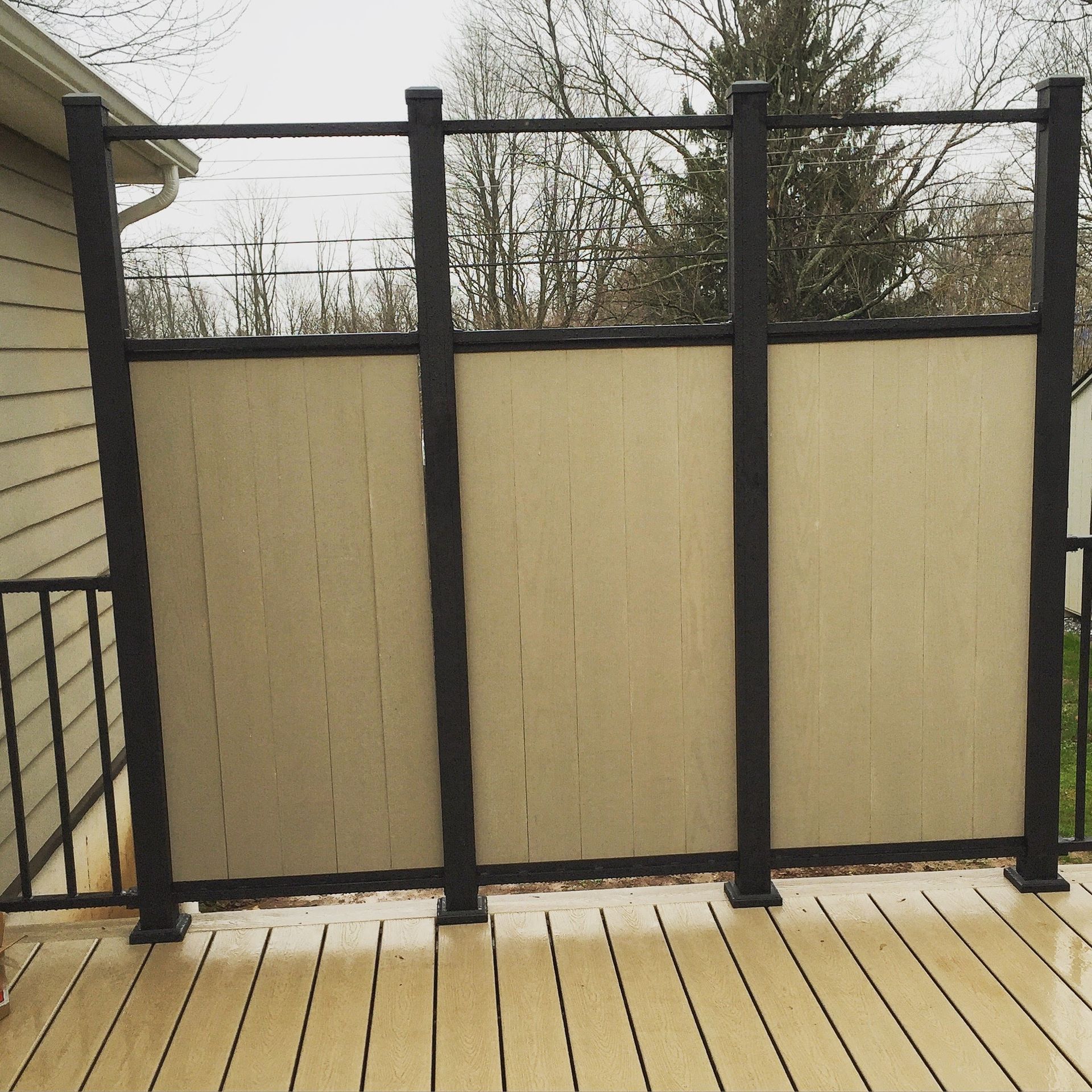 Tan and black deck privacy screen. Beige wood-like panels between black posts on a wooden deck.