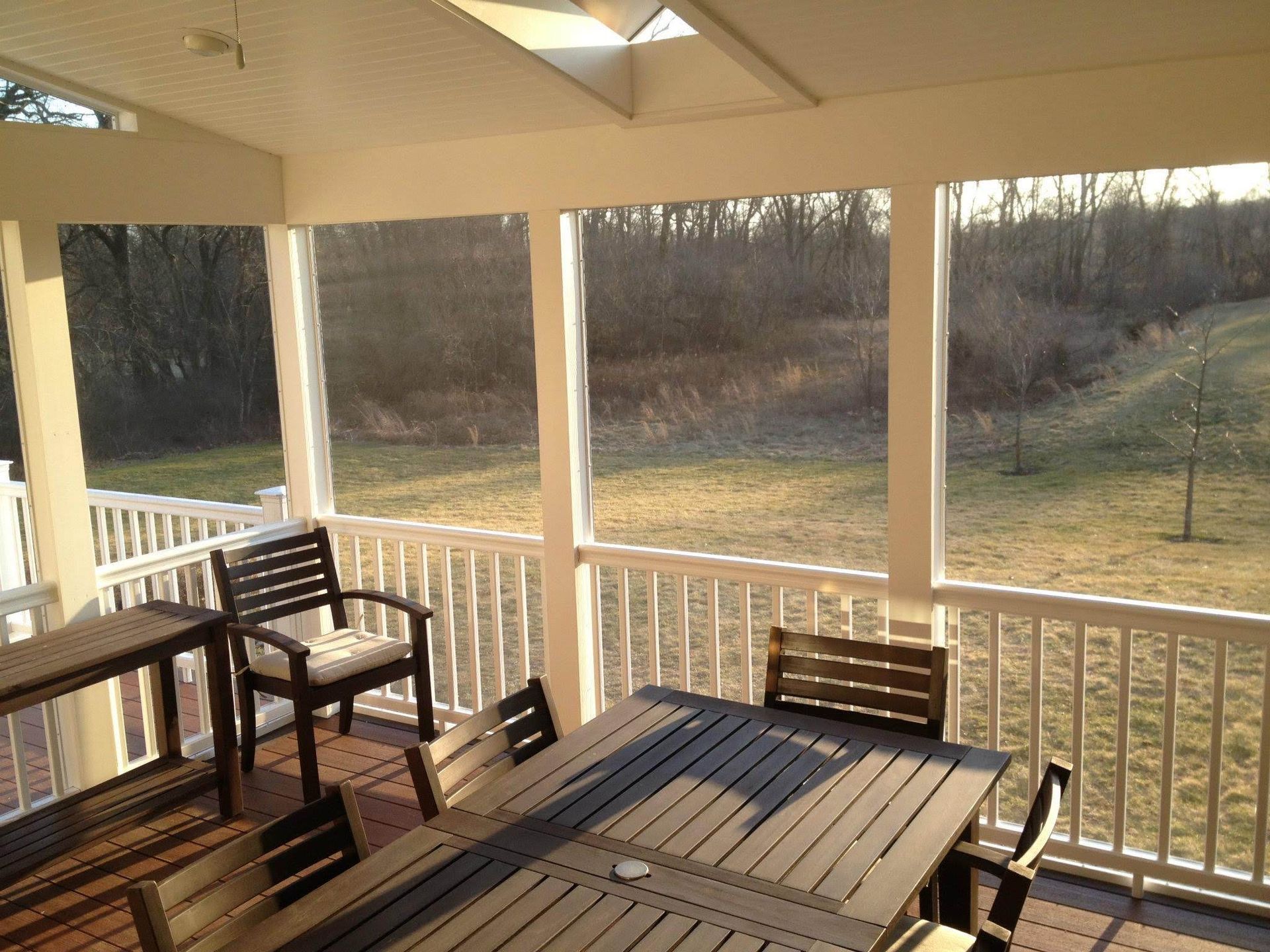 Screened-in porch with outdoor dining table and chairs, overlooking a grassy yard and trees.