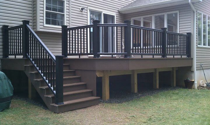 A raised brown composite deck with black metal railings and a staircase, attached to the back of a beige siding house.