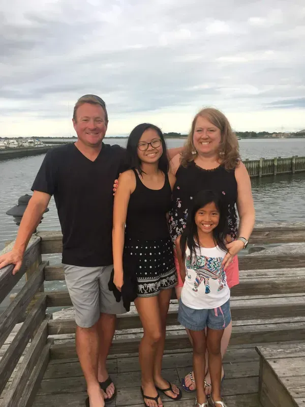 Family of four poses on a wooden pier by the water under a cloudy sky.