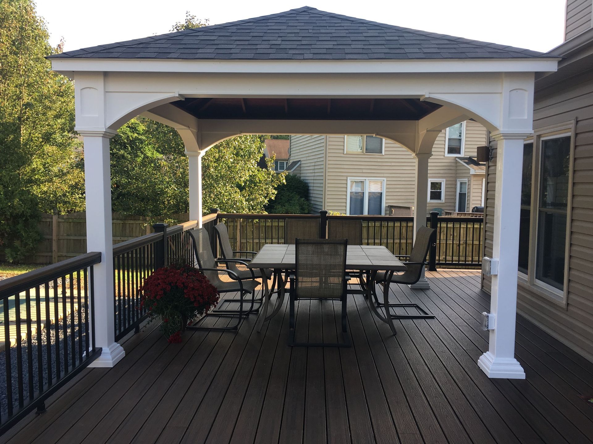 Gazebo over a wooden deck with a table and chairs, near a house with windows.