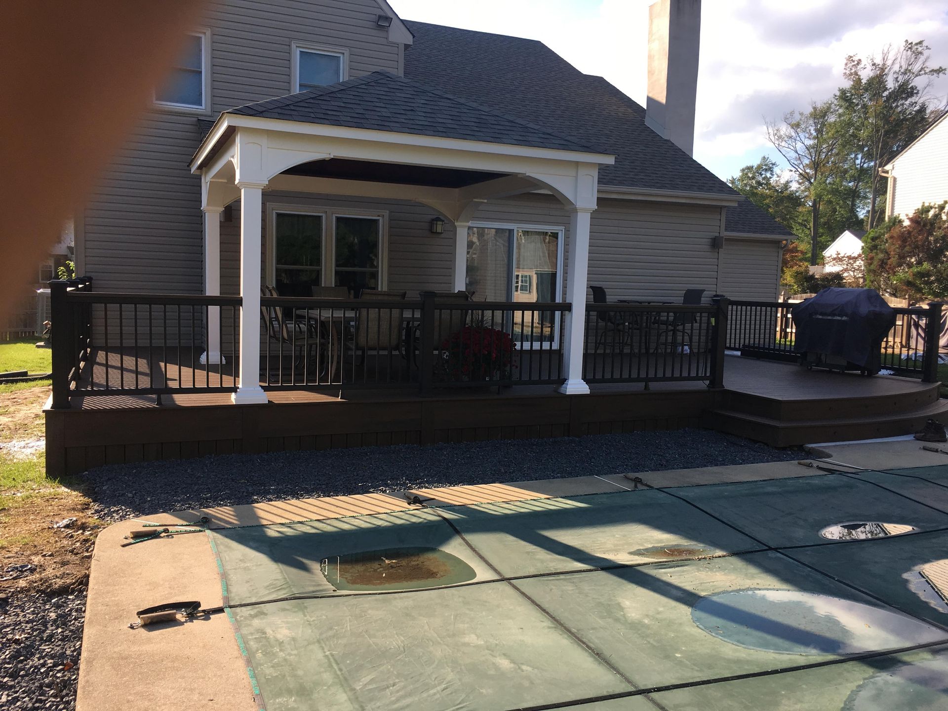 Backyard deck with a covered seating area, overlooking a pool. Brown deck, white columns, black railing.