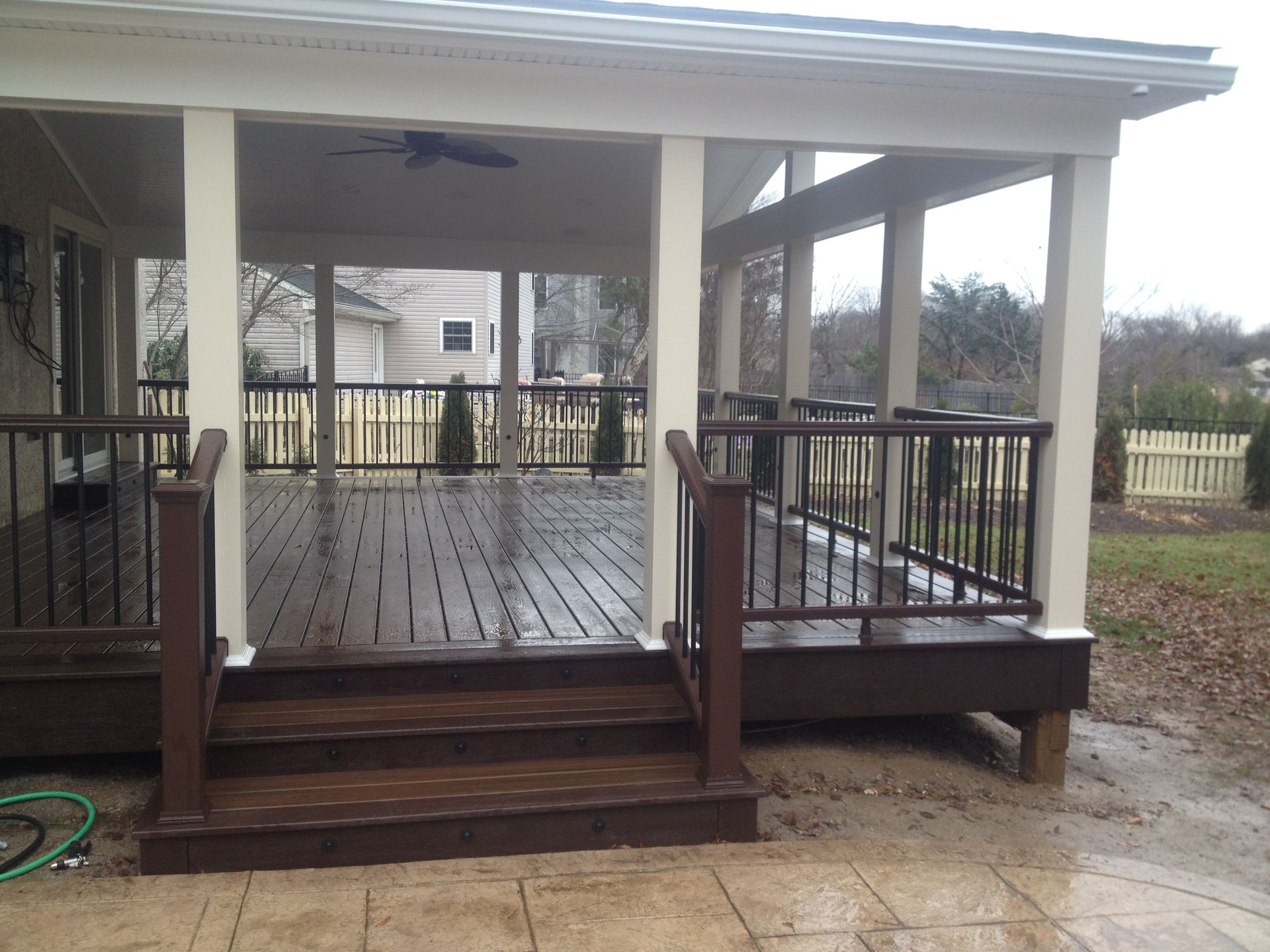 Covered wooden deck with steps, brown railings, beige columns, and a view of a backyard.