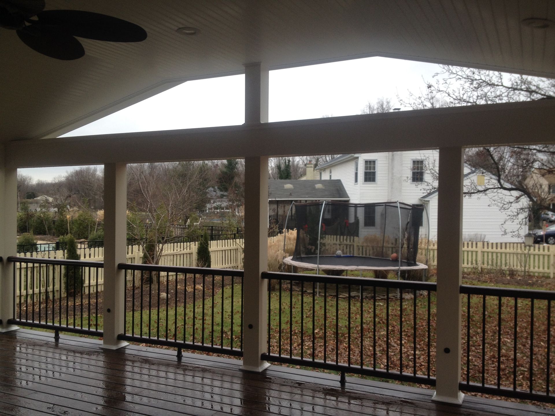 Covered deck with dark railings, supporting posts, and view of a yard with a trampoline and houses.