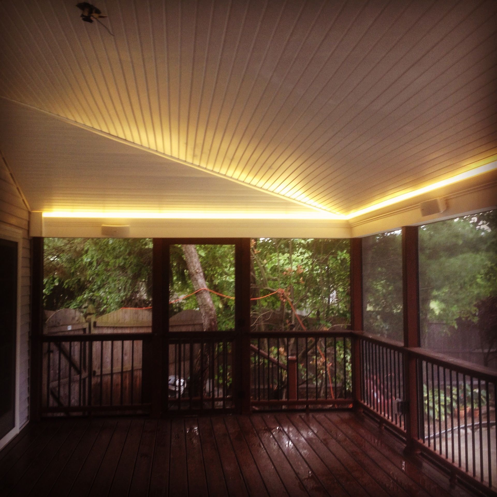 Screened porch with wooden floor and railing, with warm LED lighting on the ceiling. View of lush green trees through the screen.