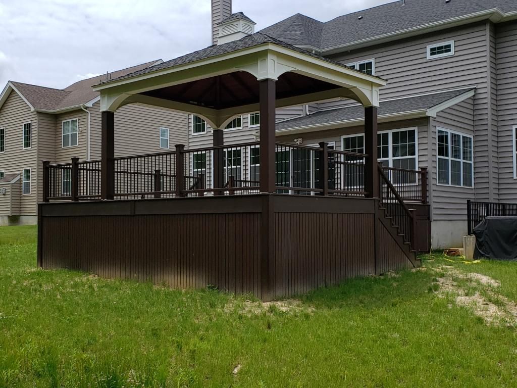Brown deck with gazebo, railing, stairs, attached to a gray house, on a grassy lawn.