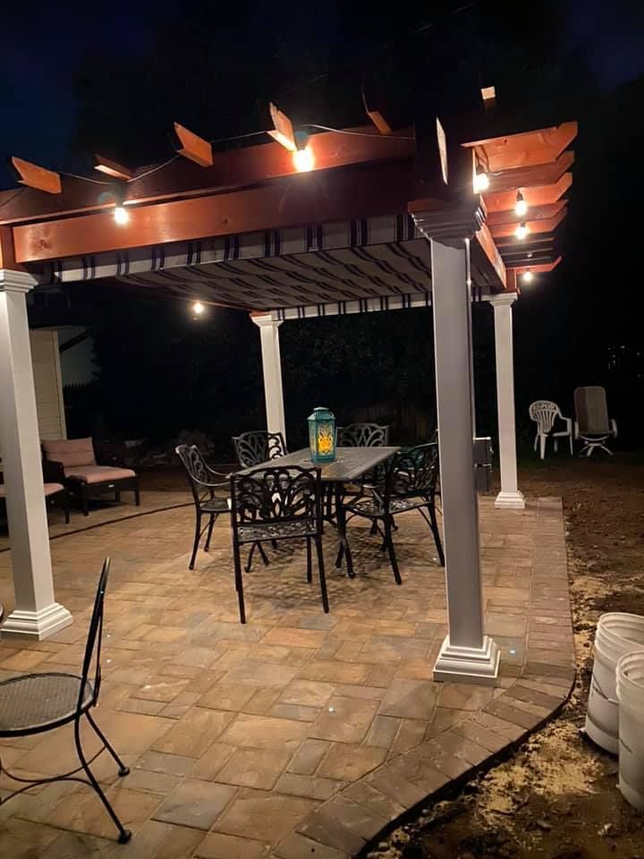 Patio with pergola, string lights, table, chairs, at night. Dark surroundings, brown pavers, white columns.