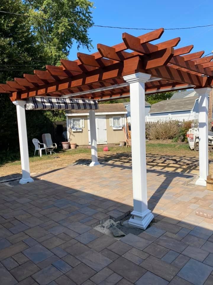 Pergola with brown wooden top and white pillars over a brick patio. Backyard setting.