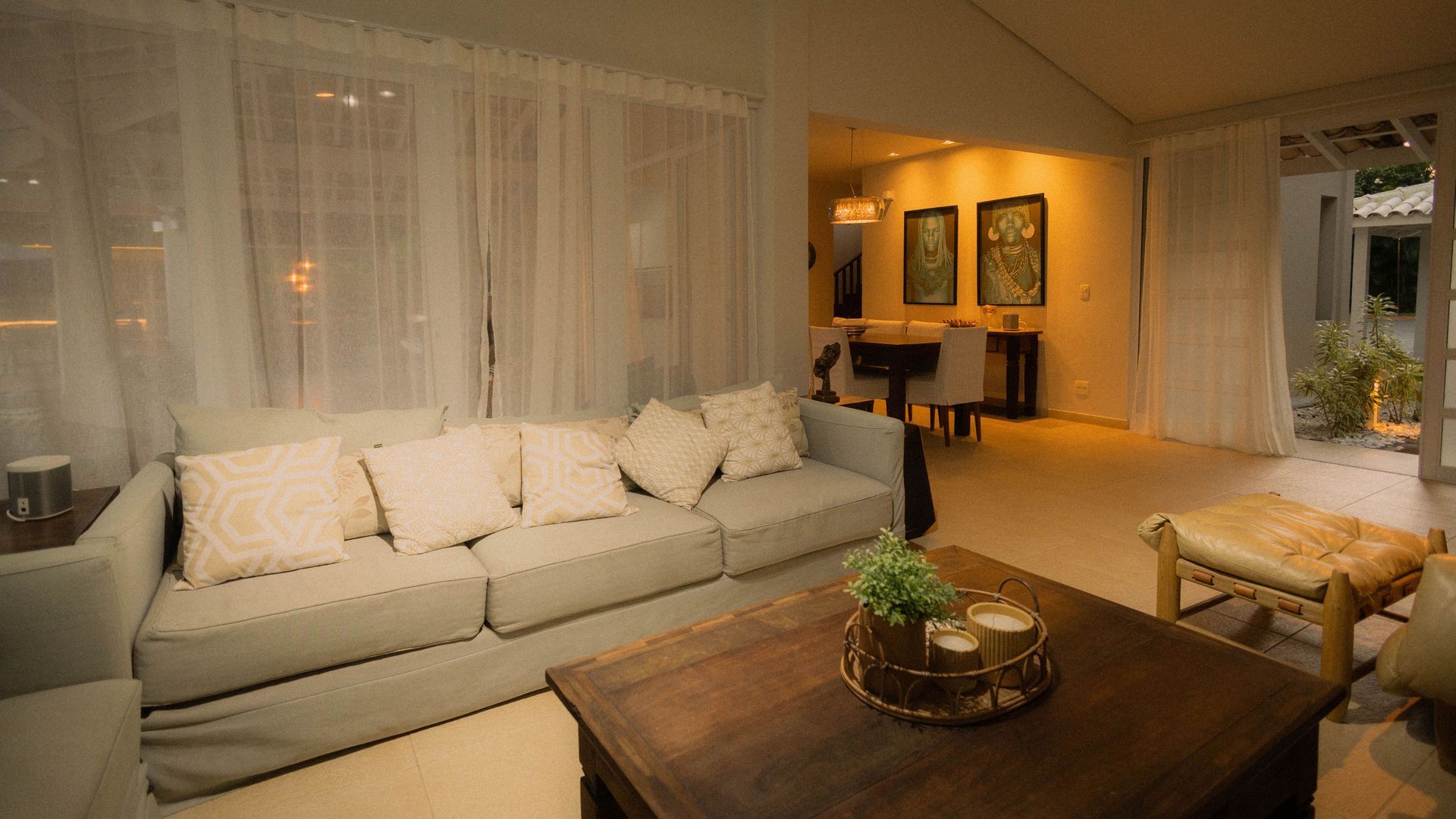 Warm living room with white sofa, wooden coffee table, sheer curtains, and softly lit dining area in back