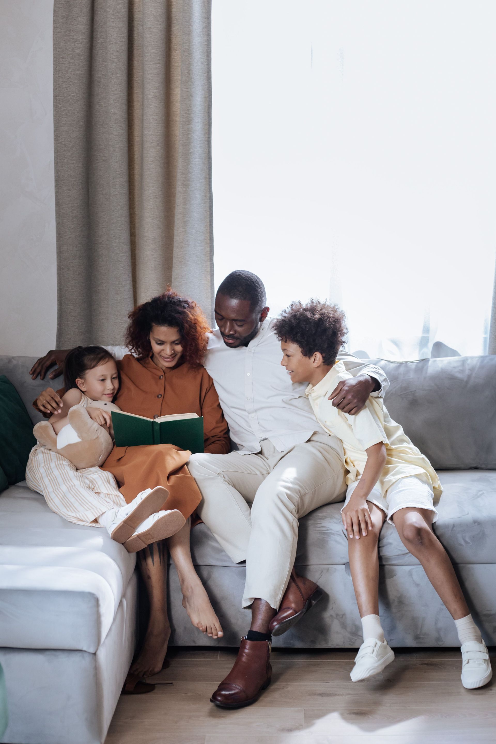 Family sitting together on a sofa, reading a book in a bright living room.