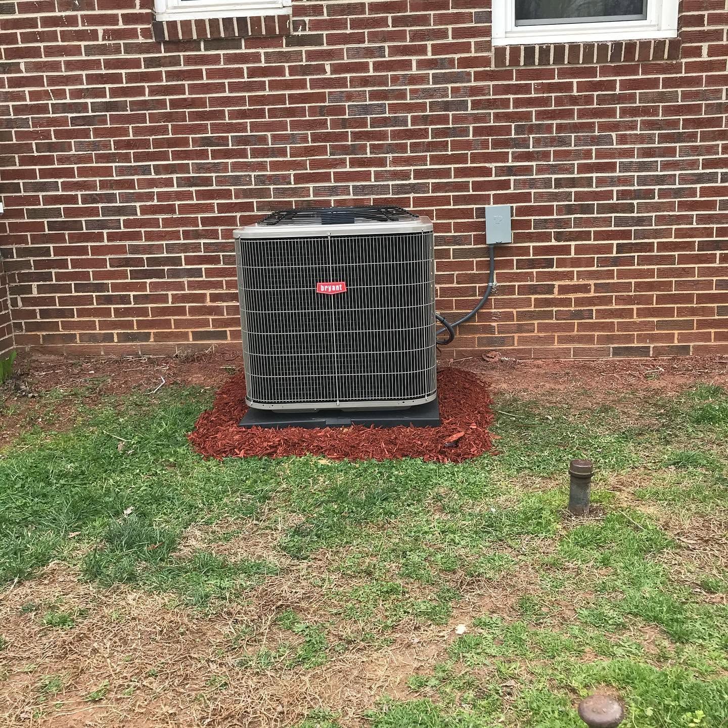 Outdoor air conditioning unit beside a brick house, with red mulch and grass in the yard.