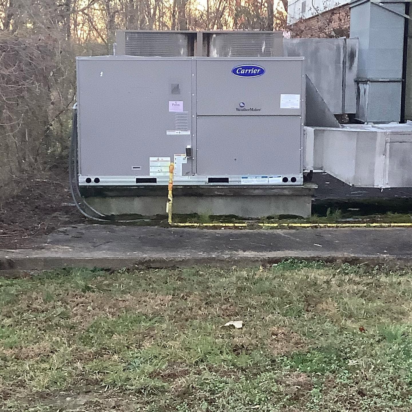 Gray commercial HVAC unit beside a building, with a yellow gas pipe and wooded yard in front.