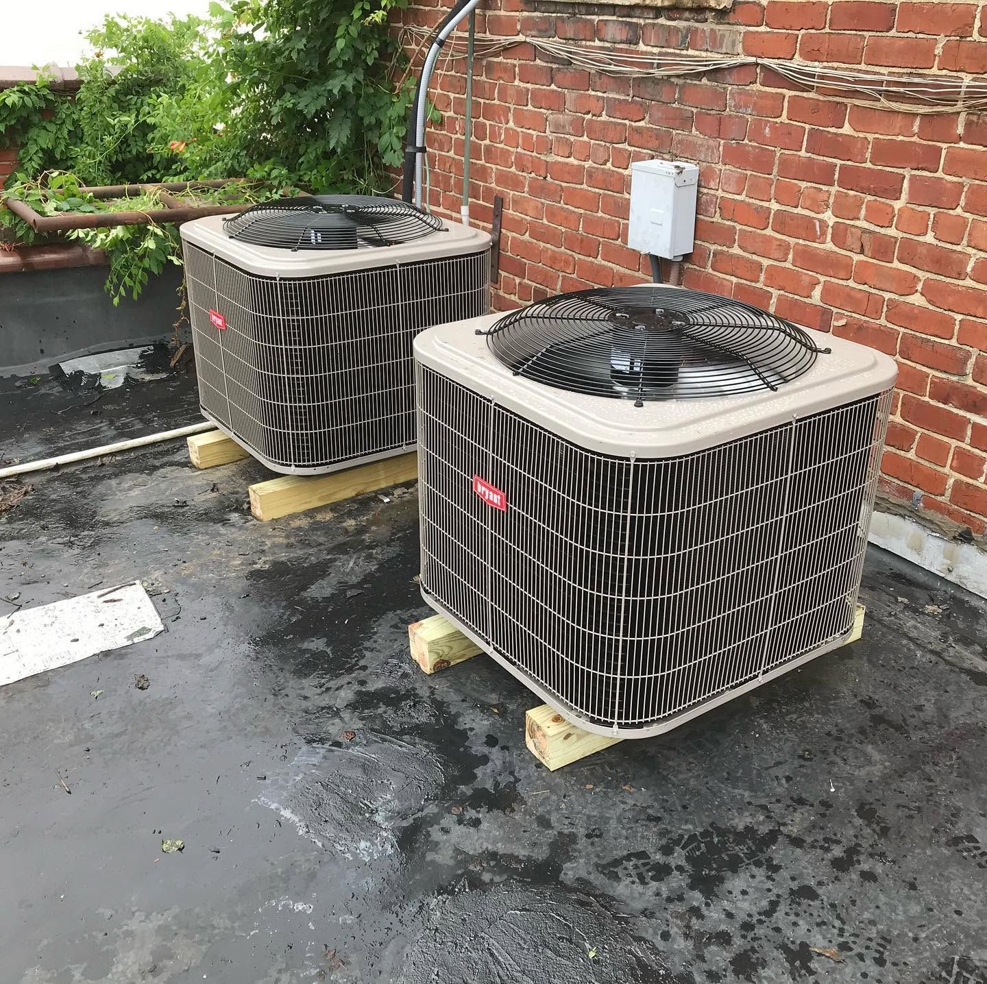 Two outdoor HVAC units on wooden blocks beside a brick wall on a wet rooftop.